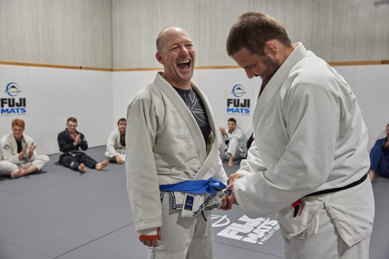 Two men in Judo uniforms with one receiving a blue belt in a gym setting.